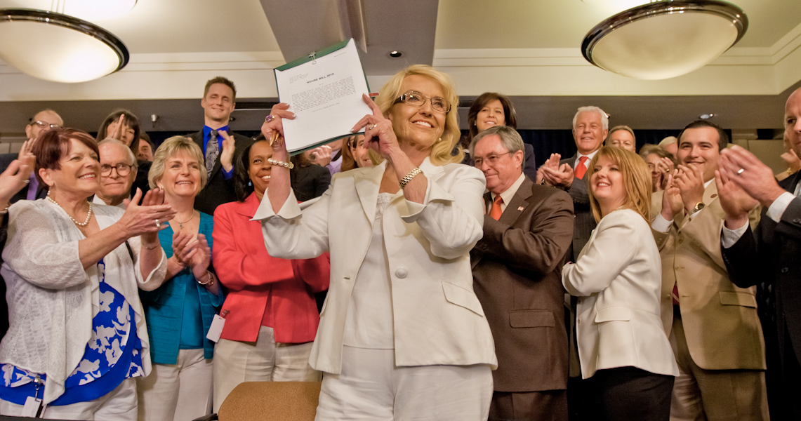 Gov. Jan Brewer holds the newly signed Medicaid expansion bill as the bipartisan coalition of supporters who helped pass the bill applaud at the June 17 bill signing. (Photo by Evan Wyloge/Arizona Capitol Times)