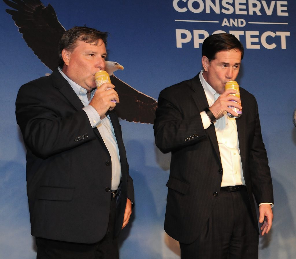 Gov. Doug Ducey, right, sips on a cold one during a ceremony announcing a a Flagstaff craft brewery will link donations to the state Game and Fish Department to sales of one of its lines of beer. With him is Kurt Davis of the Game and Fish Commission. (Capitol Media Services photo by Howard Fischer)